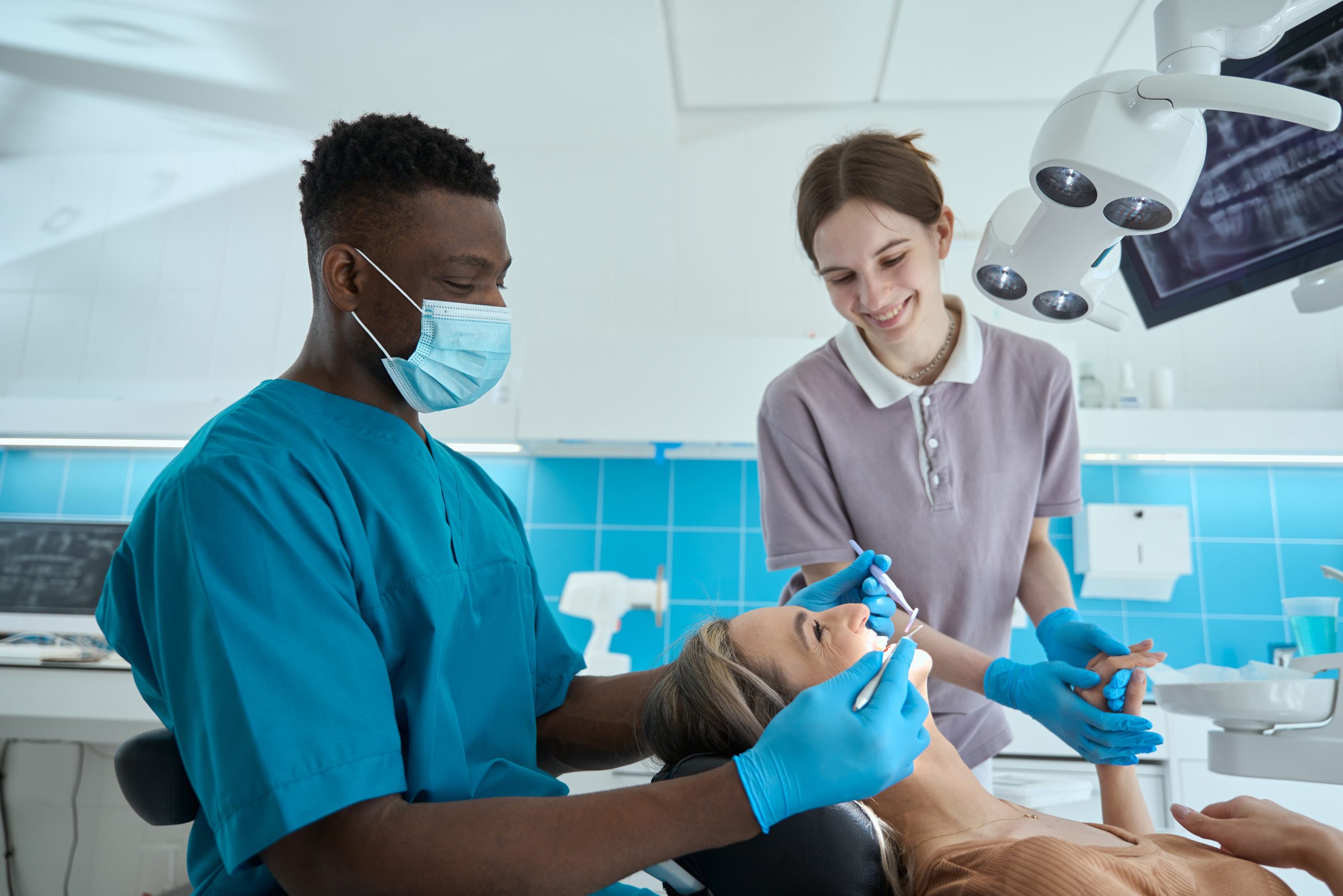 Médecin en masque de protection et costume bleu assis sur une chaise près du patient et tenant un miroir dentaire<br />
