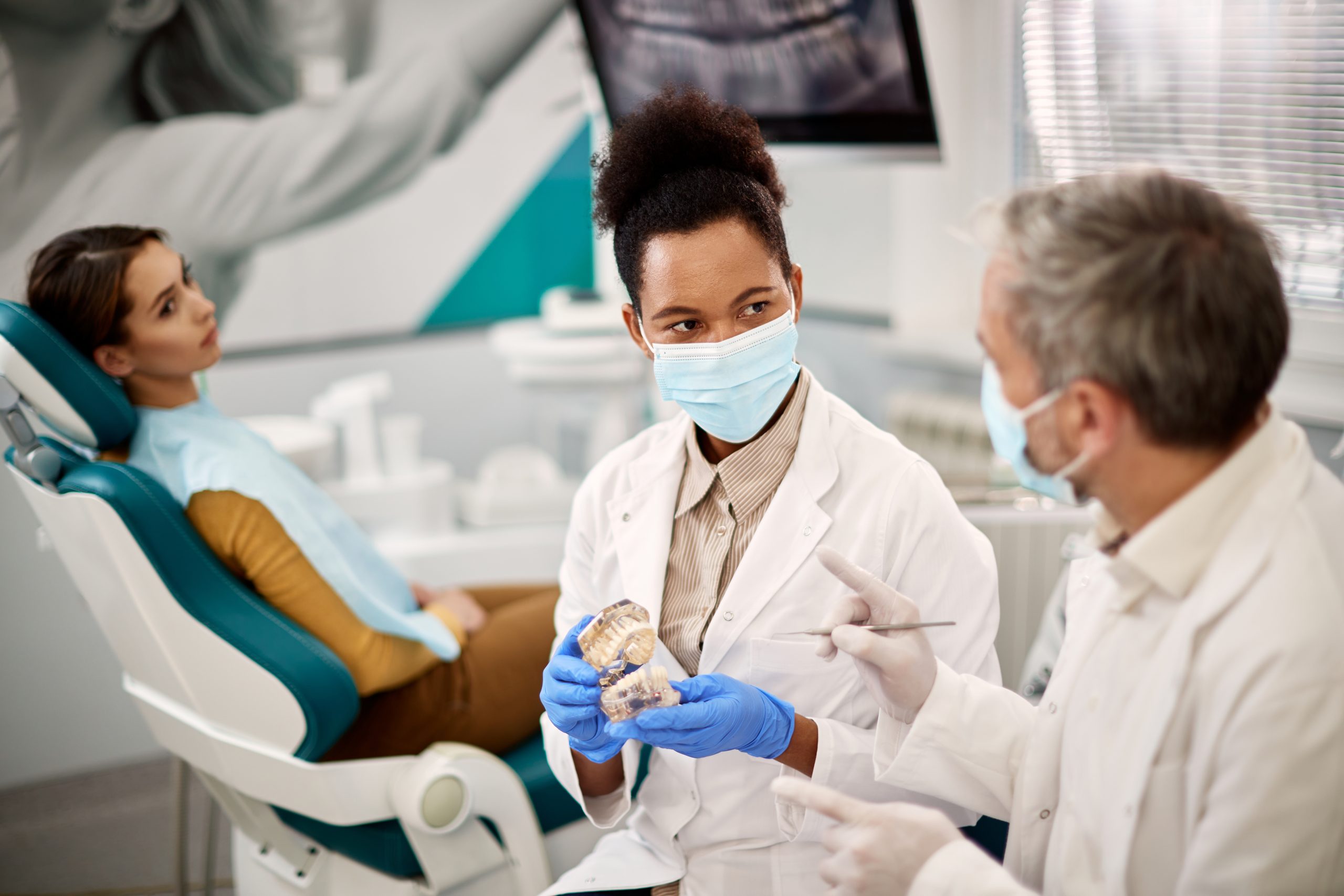 Dentiste afro-américain utilisant des prothèses dentaires et parlant à son collègue tout en ayant rendez-vous avec une patiente à la clinique dentaire.